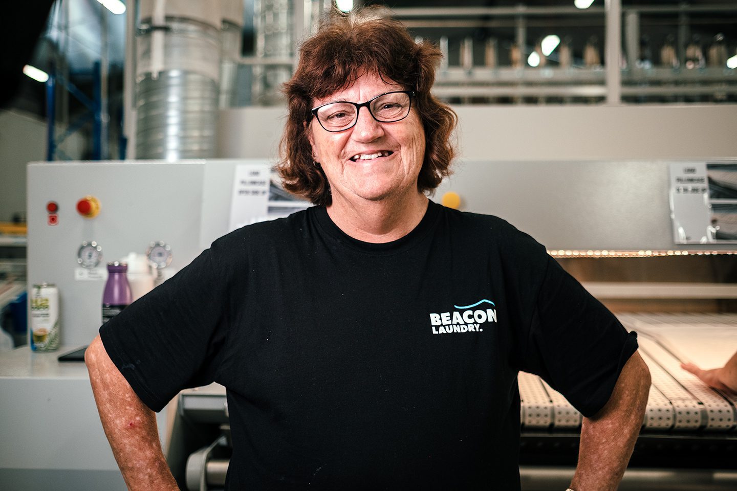Leigh, a Beacon Laundry employee, standing in front of laundry machinery smiling.