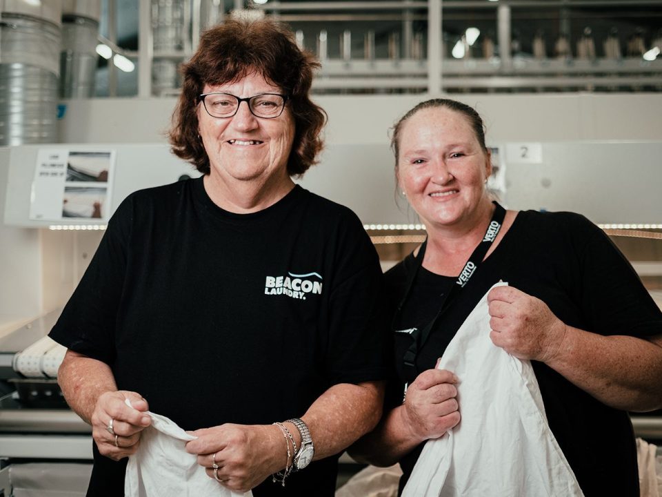 Two Beacon Laundry employees standing side by side smiling, holding sheets in a laundry.
