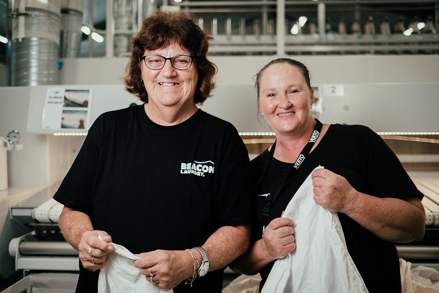 Two Beacon Laundry employees standing side by side smiling, holding sheets in a laundry.