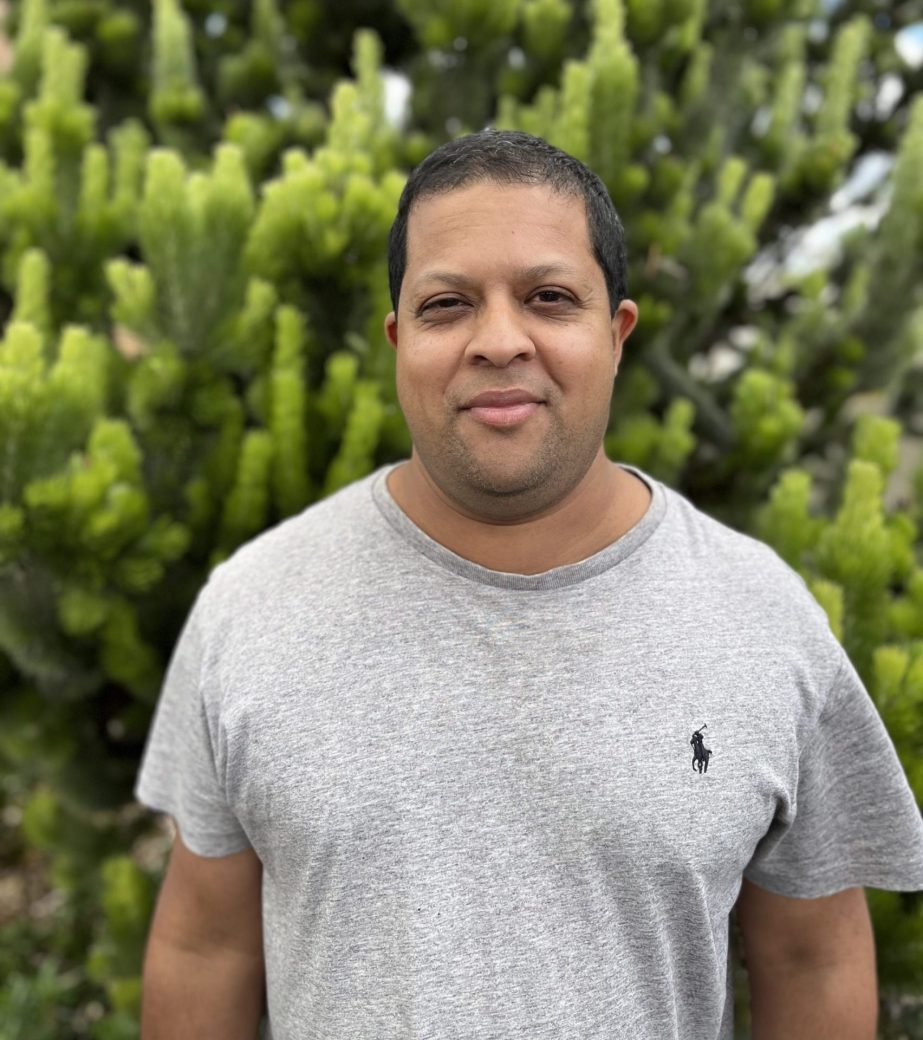 Headshot of Dominique Loulie. Man in grey t-shirt standing in front of greenery.